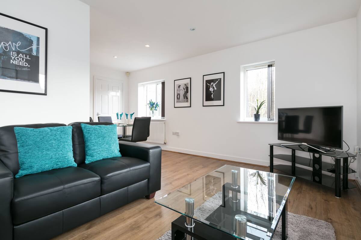 A modern living room with a black leather sofa, teal cushions, a glass coffee table, a flat-screen TV, and framed artwork on the walls.
