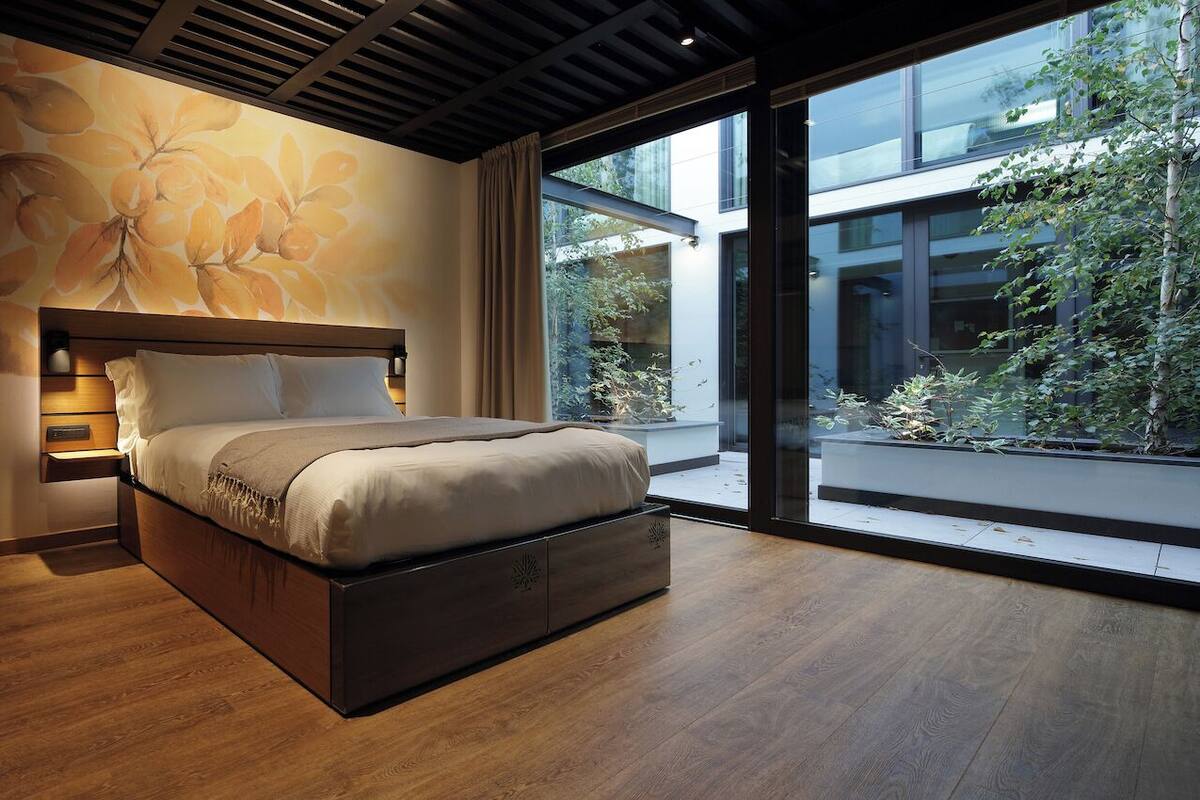 A modern bedroom with a large bed, wooden floor, and a view of greenery outside.