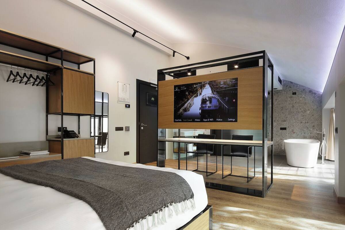 A modern hotel room with a bed, a TV mounted on a wooden panel, a desk, and a large window.