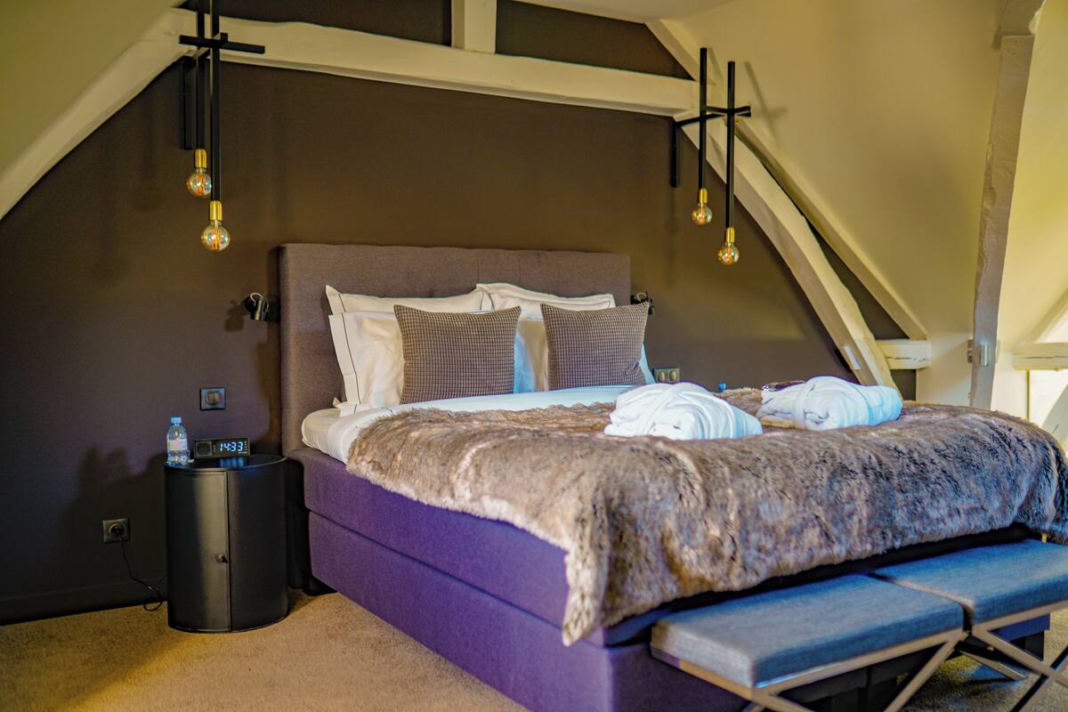 A bedroom with a purple bed, a bench, and a nightstand with a water bottle and clock.