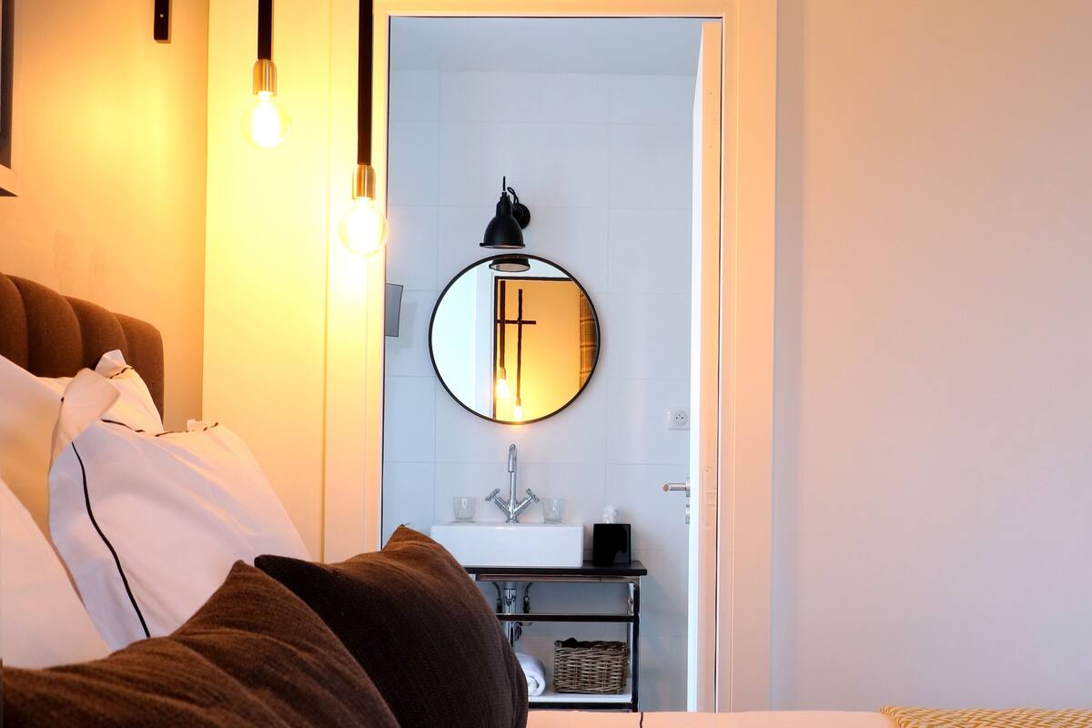 A bedroom with a bed, pillows, a bedside table, a mirror, and a door leading to a bathroom.