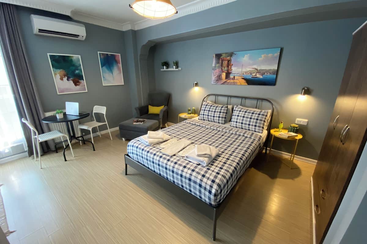 A neatly arranged bedroom with a bed, a desk, and a chair. There is a painting on the wall and an air conditioning unit.