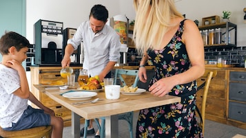 Una familia comiendo en la encimera de la cocina, con una cafetera, fruta y pasteles.
