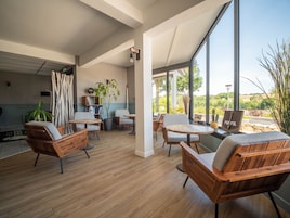 Un interior moderno con suelo de madera, una zona de estar con sofás y una mesa, una estantería y un amplio ventanal con vistas a zonas verdes.