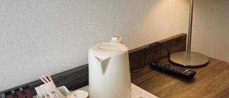 A hotel room with a bedside table featuring a lamp, a remote control, and a tray with coffee cups and tea packets.