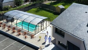 A covered swimming pool with a glass enclosure, surrounded by a garden with plants and a fenced area. - Hotel du Trégor (Treguier)