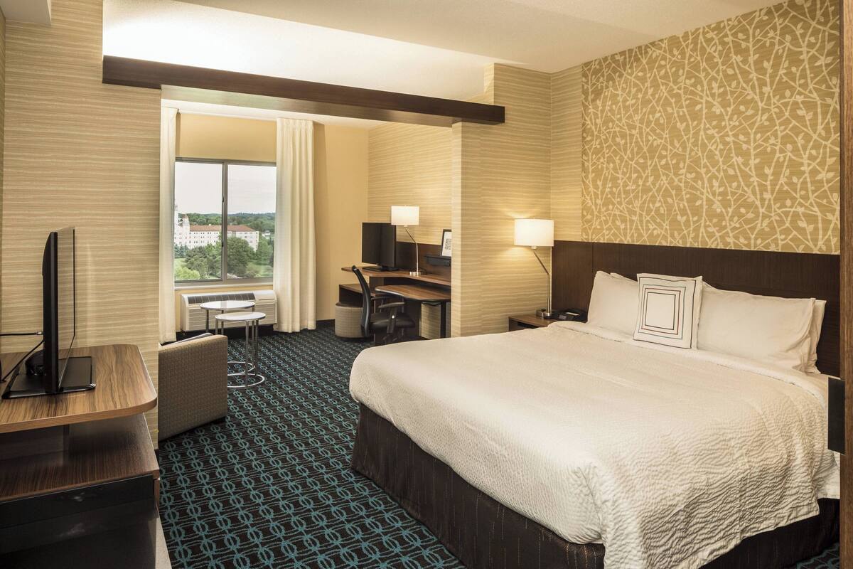 Habitación de hotel con una cama grande, un escritorio, una silla, un televisor y vistas a la ciudad.