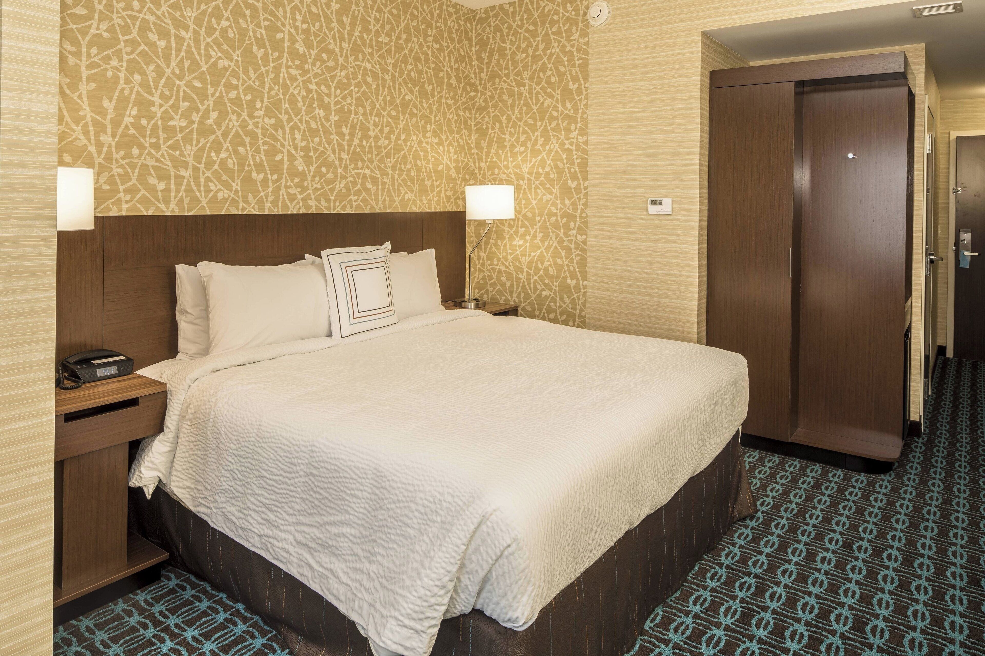 Habitación de hotel con una cama grande, una mesita de noche con lámpara, un armario de madera y una alfombra estampada.