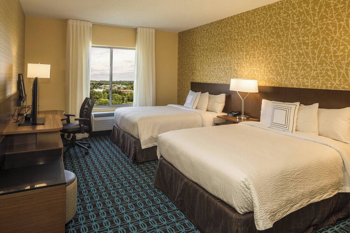 Habitación de hotel con dos camas, un escritorio, una silla y vistas al exterior.