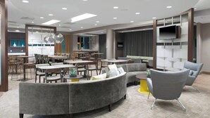 A modern hotel lobby with a curved grey sofa, round tables, and a flat-screen TV mounted on a wall. - SpringHill Suites by Marriott Kansas City Lenexa City Center (Lenexa)
