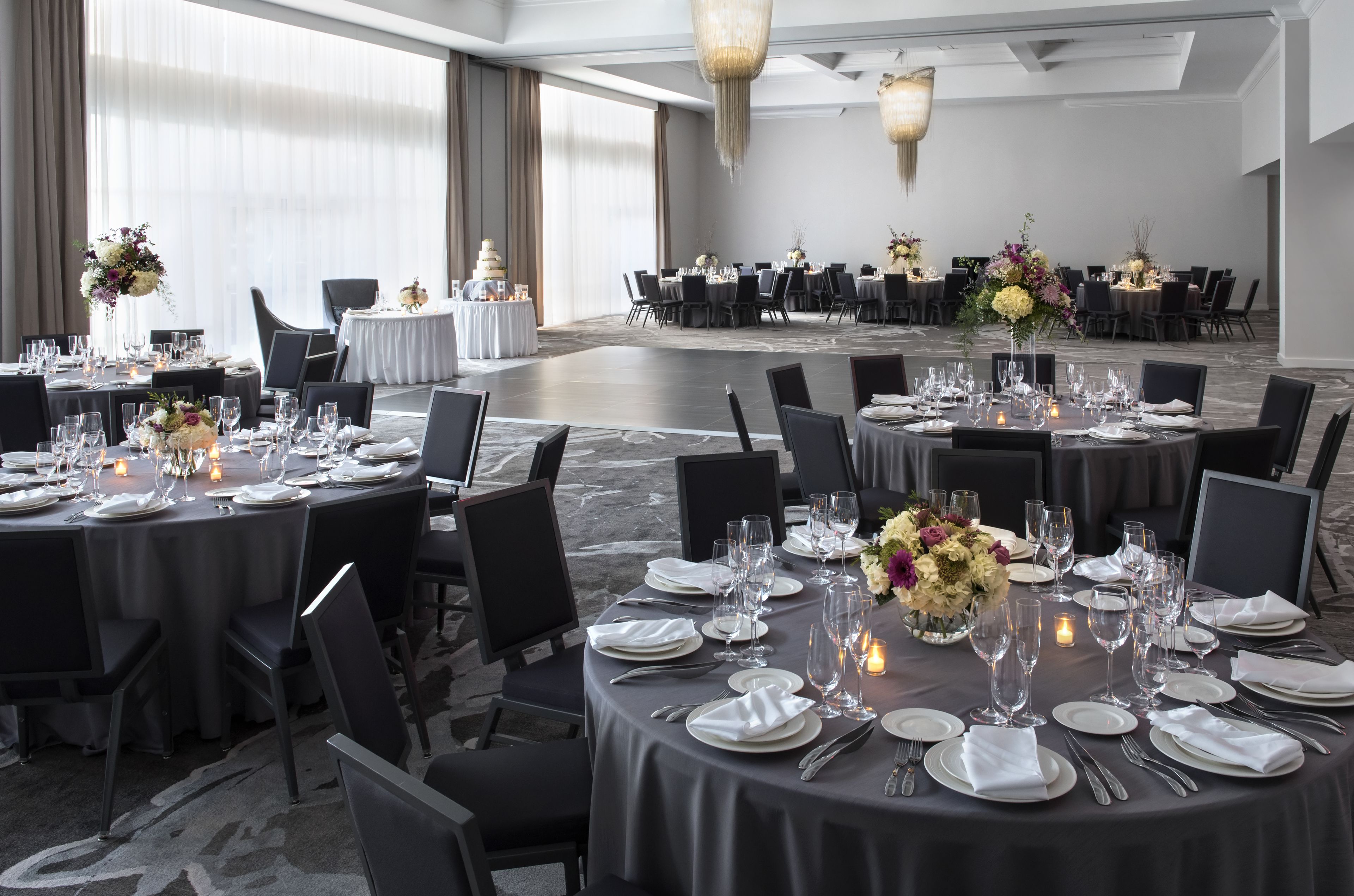 Ein Festsaal mit runden Tischen, gedeckt für ein Essen, mit weißen Tischdecken, schwarzen Stühlen und floralen Mittelstücken.
