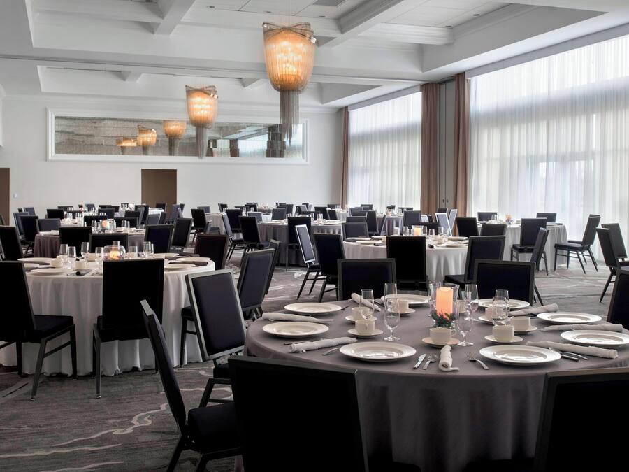 Un gran saló de banquets amb taules rodones preparades per dinar, amb lluminàries modernes i grans finestres amb cortines.
