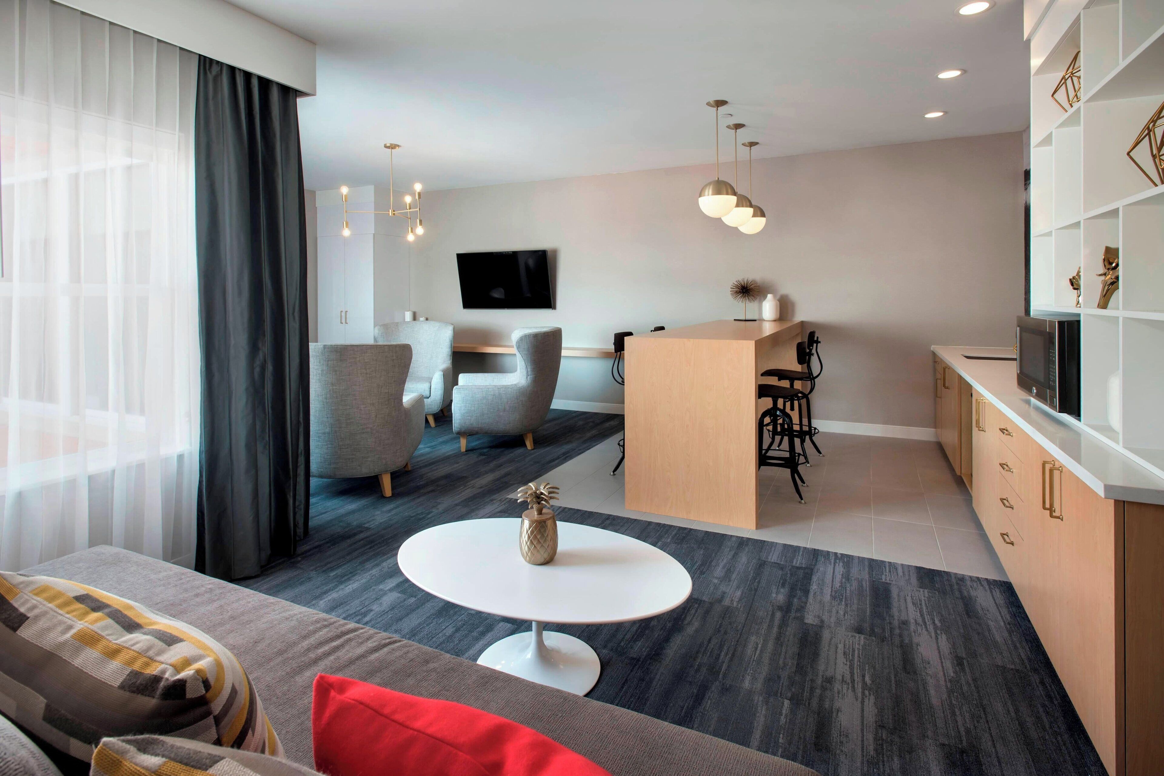 A modern living room with a sofa, a round coffee table, and a TV mounted on the wall. There is a kitchen area with a microwave and a bar counter.