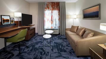 Habitación de hotel con televisión de pantalla plana, un sofá, un escritorio con silla, una mesita y una ventana con cortinas.