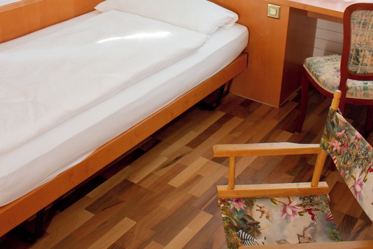 Uma cama de solteiro com cabeceira de madeira, uma escrivaninha com telefone, uma cadeira com padrão floral e um quadro na parede.