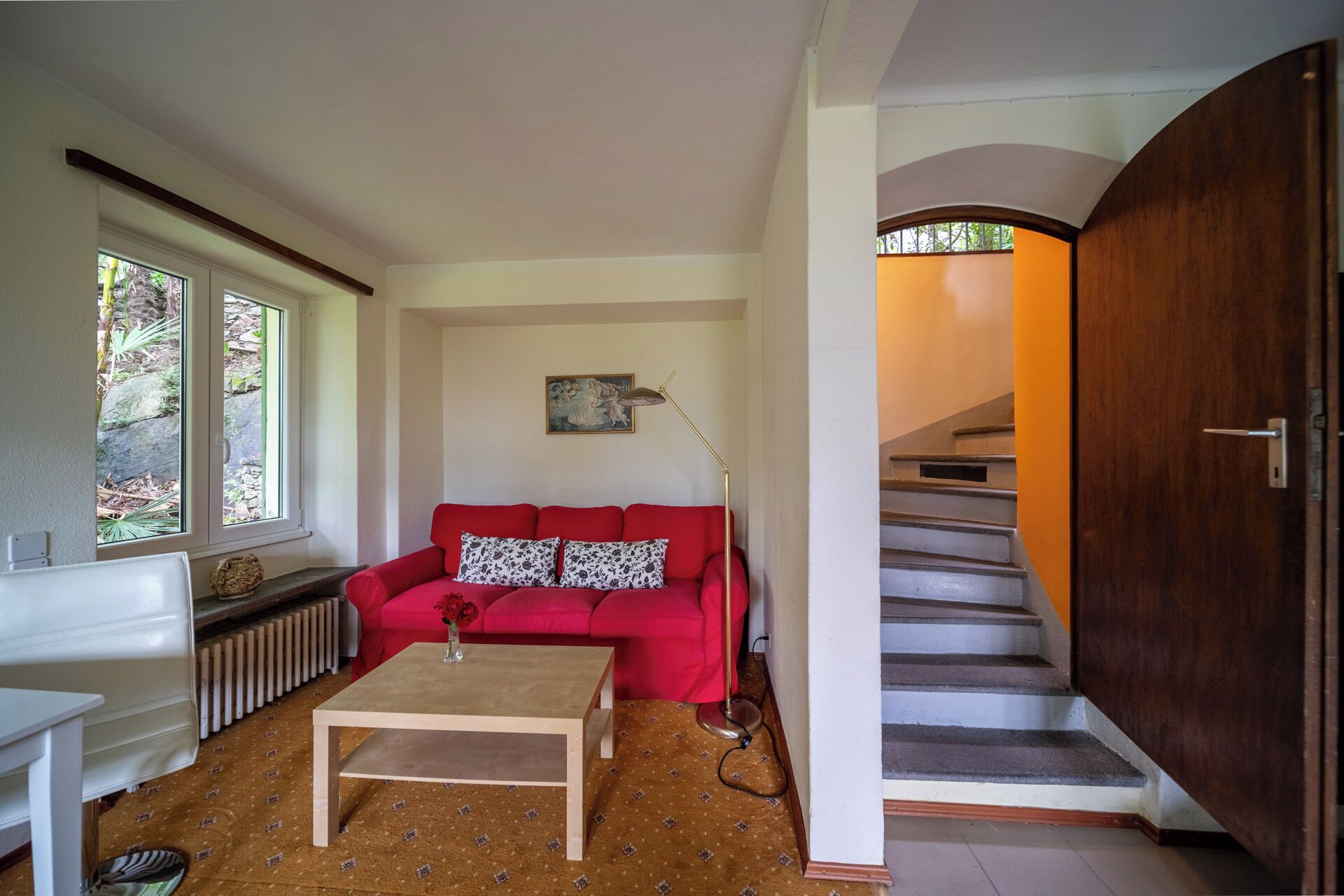 Sala de estar com sofá vermelho, mesa de centro e radiador. Escada com porta de madeira que dá acesso a outro cômodo.