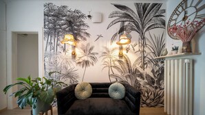 Ein Wohnzimmer mit einem dunkelblauen Samtsofa, zwei Wandleuchten und einem tropischen Wandgemälde.