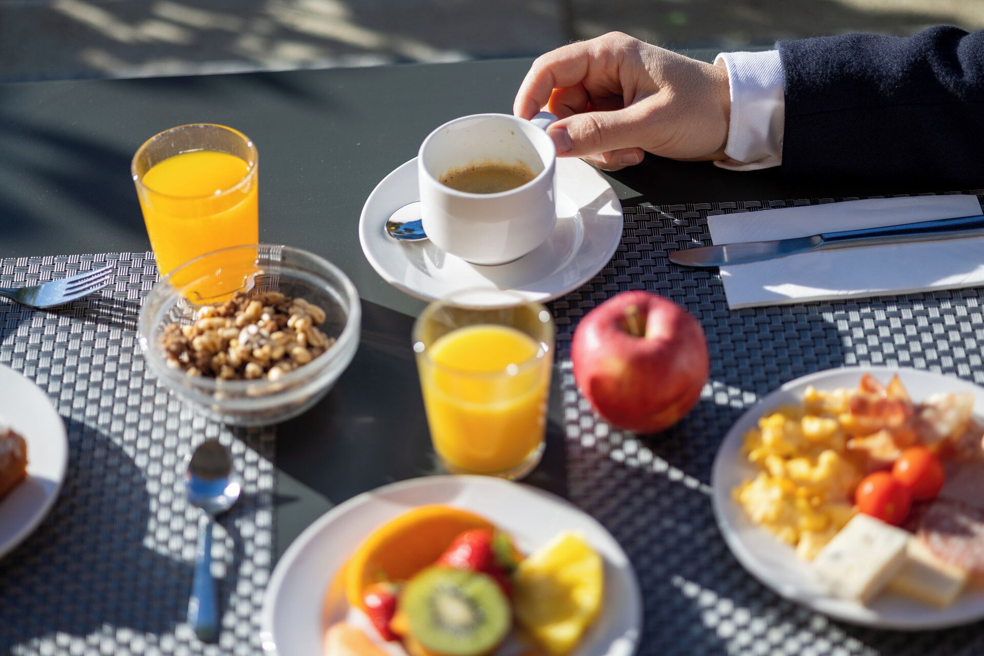 Et morgenbord med en kop kaffe, appelsinjuice, en skål müsli, et æble og en tallerken med frugt og ost.