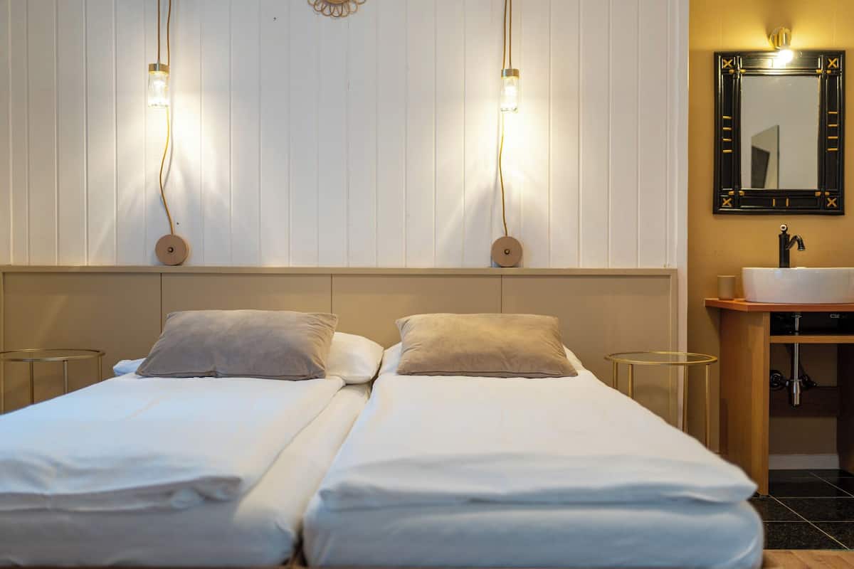 Quarto de hotel com duas camas, uma mesinha de cabeceira, um espelho e uma pia.