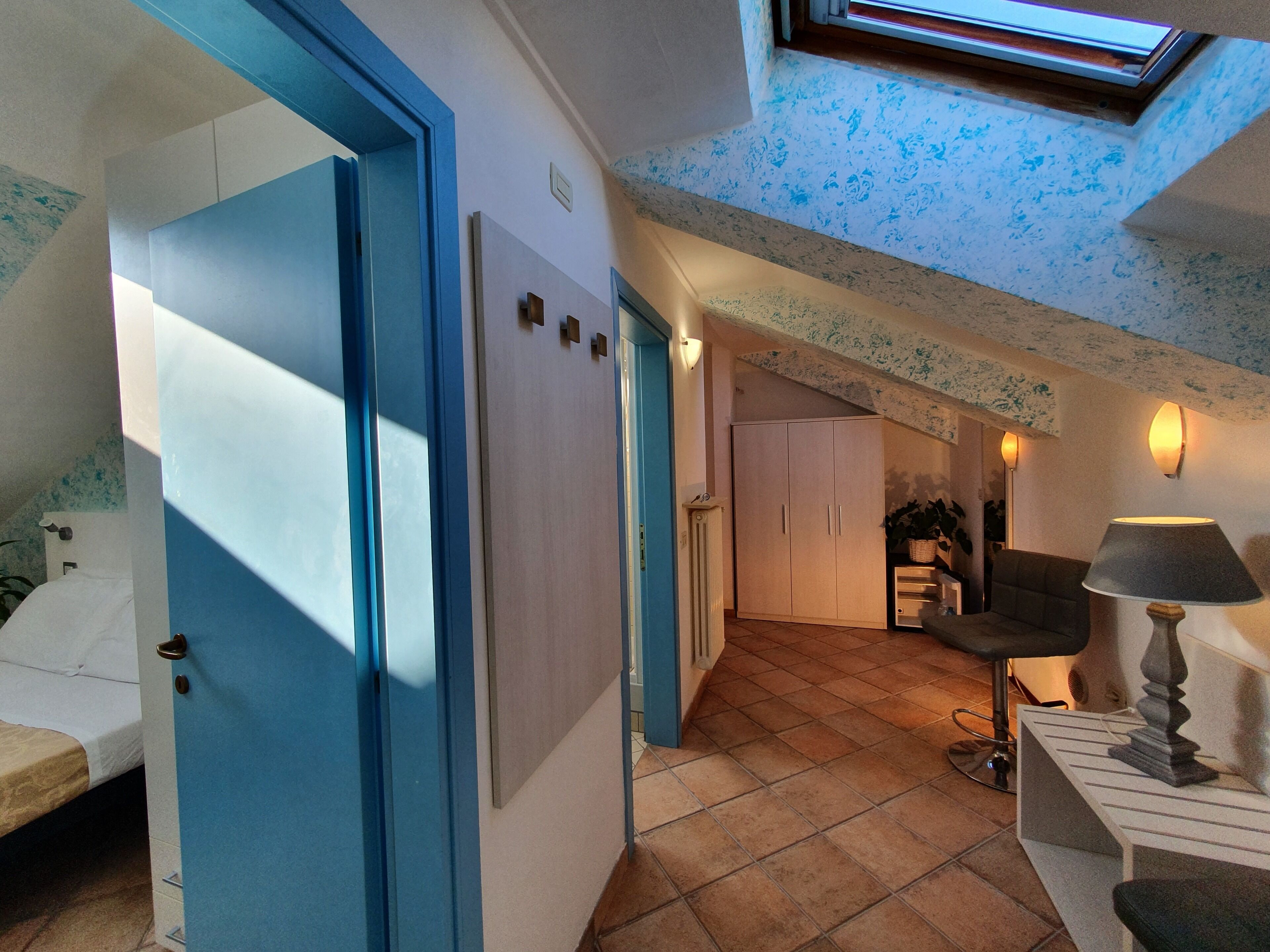 A room with a bed, a desk, a chair, and a lamp. There is a door leading to another room.