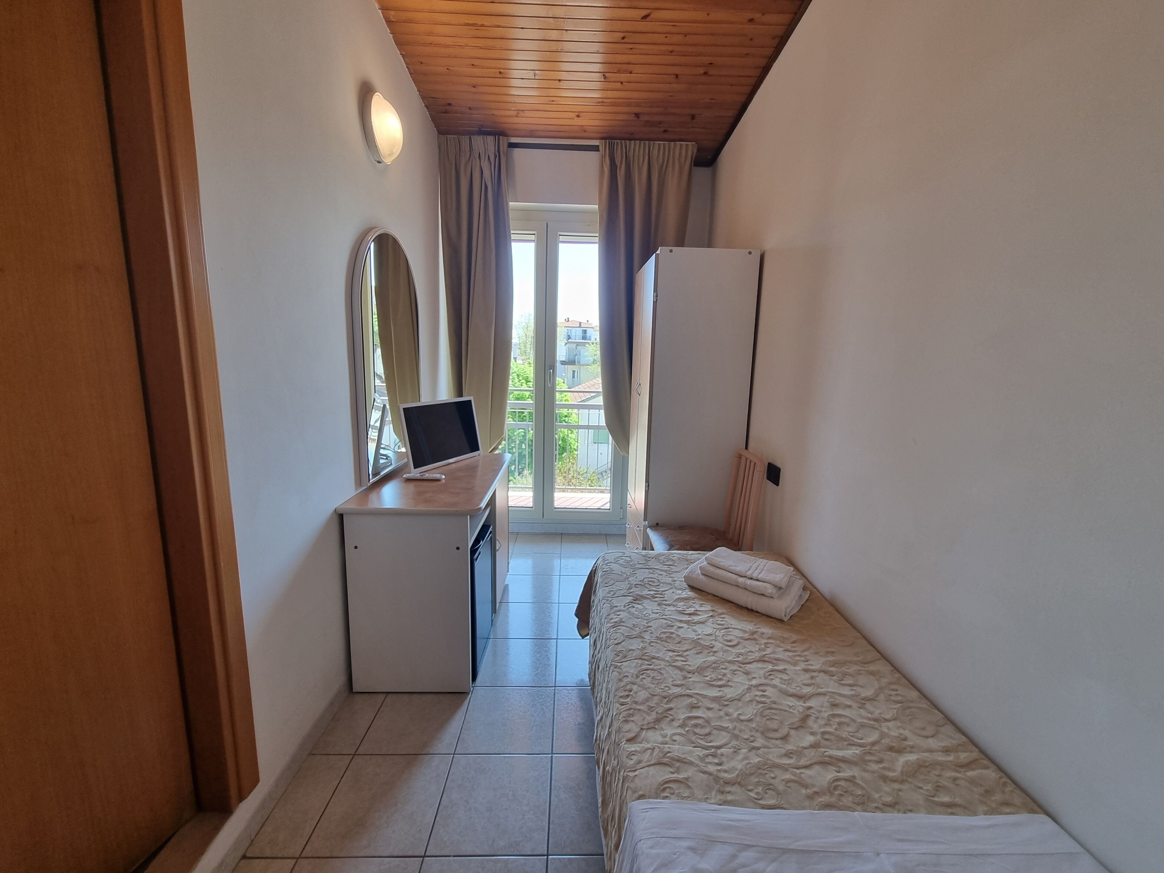 A small, well-lit bedroom with a bed, a desk with a laptop, a mirror, and a wardrobe.