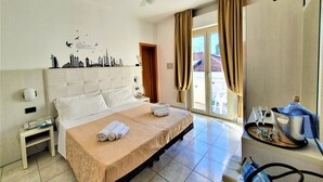 Une chambre d’hôtel avec un grand lit, un ventilateur de plafond, une décoration murale représentant un paysage urbain, une petite table avec une bouilloire et une vue sur l’extérieur grâce à une porte coulissante.