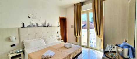 A hotel room with a large bed, a ceiling fan, a cityscape wall decoration, a small table with a kettle, and a view of the outside through a sliding door.