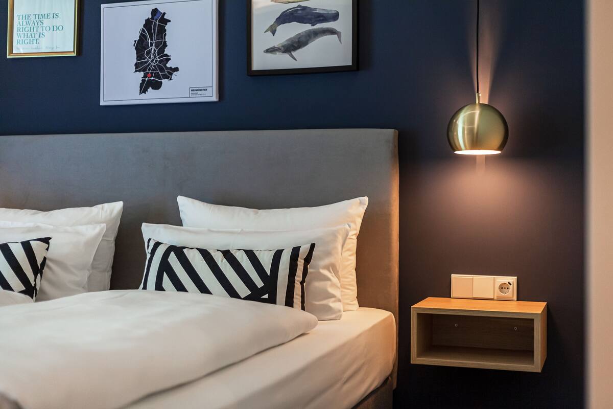 Una cama bien hecha con almohadas a rayas blancas y negras, una mesita de noche fijada a la pared con una lámpara y cuadros colgados sobre la cama.