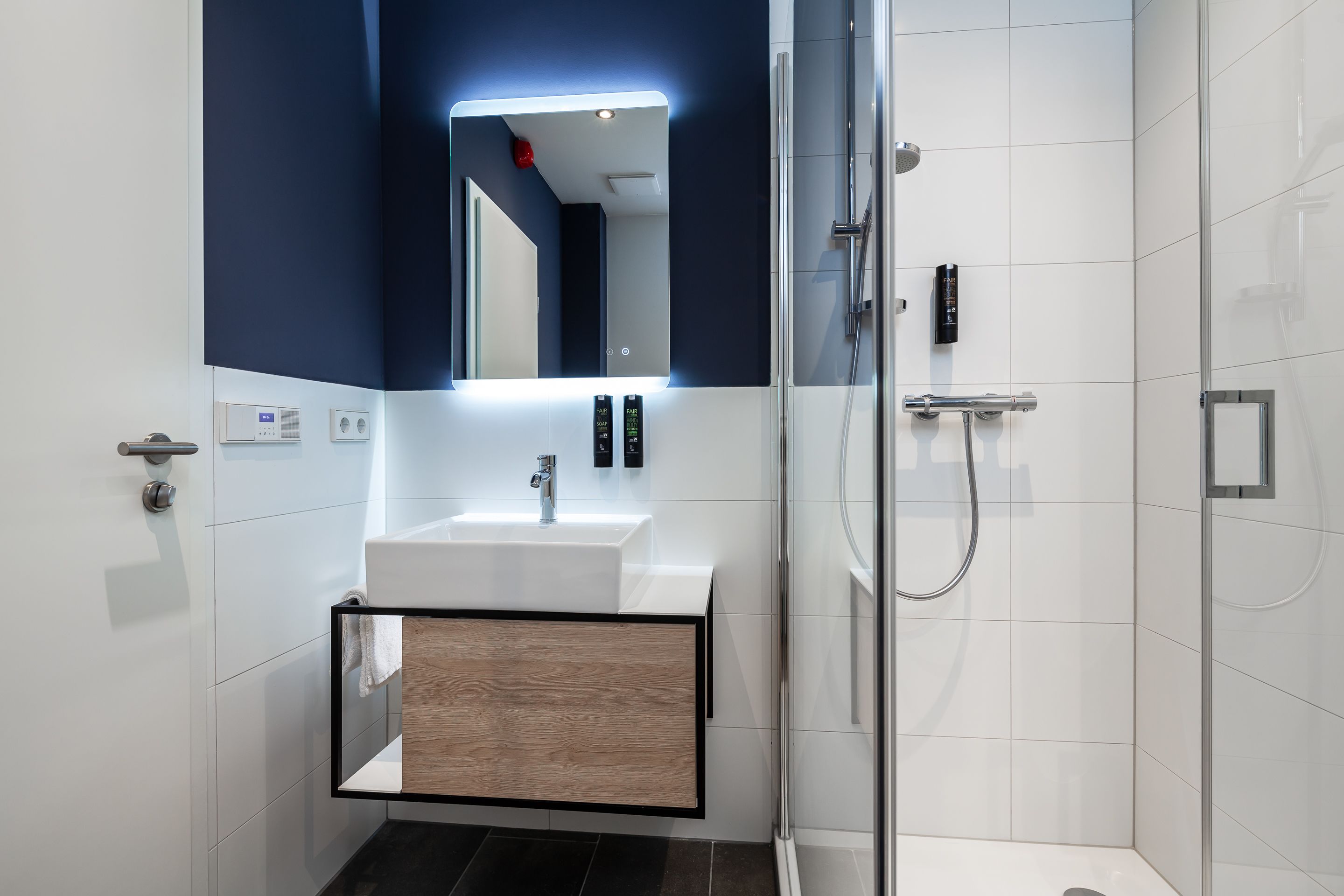 A modern bathroom with a white sink, a mirror with integrated lighting, a glass shower enclosure, and a wooden cabinet.