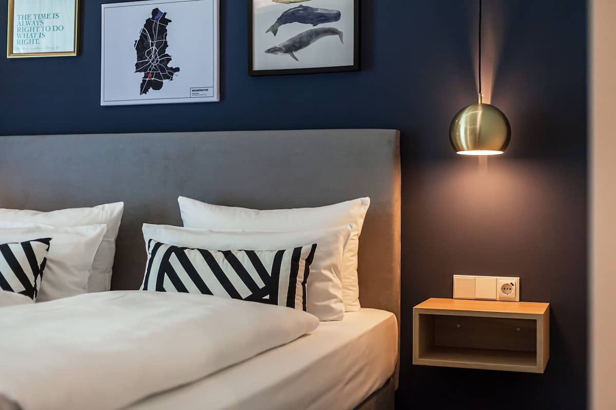 Una cama bien tendida con almohadas a rayas, una mesita de noche fijada a la pared con una lámpara y cuadros en la pared.