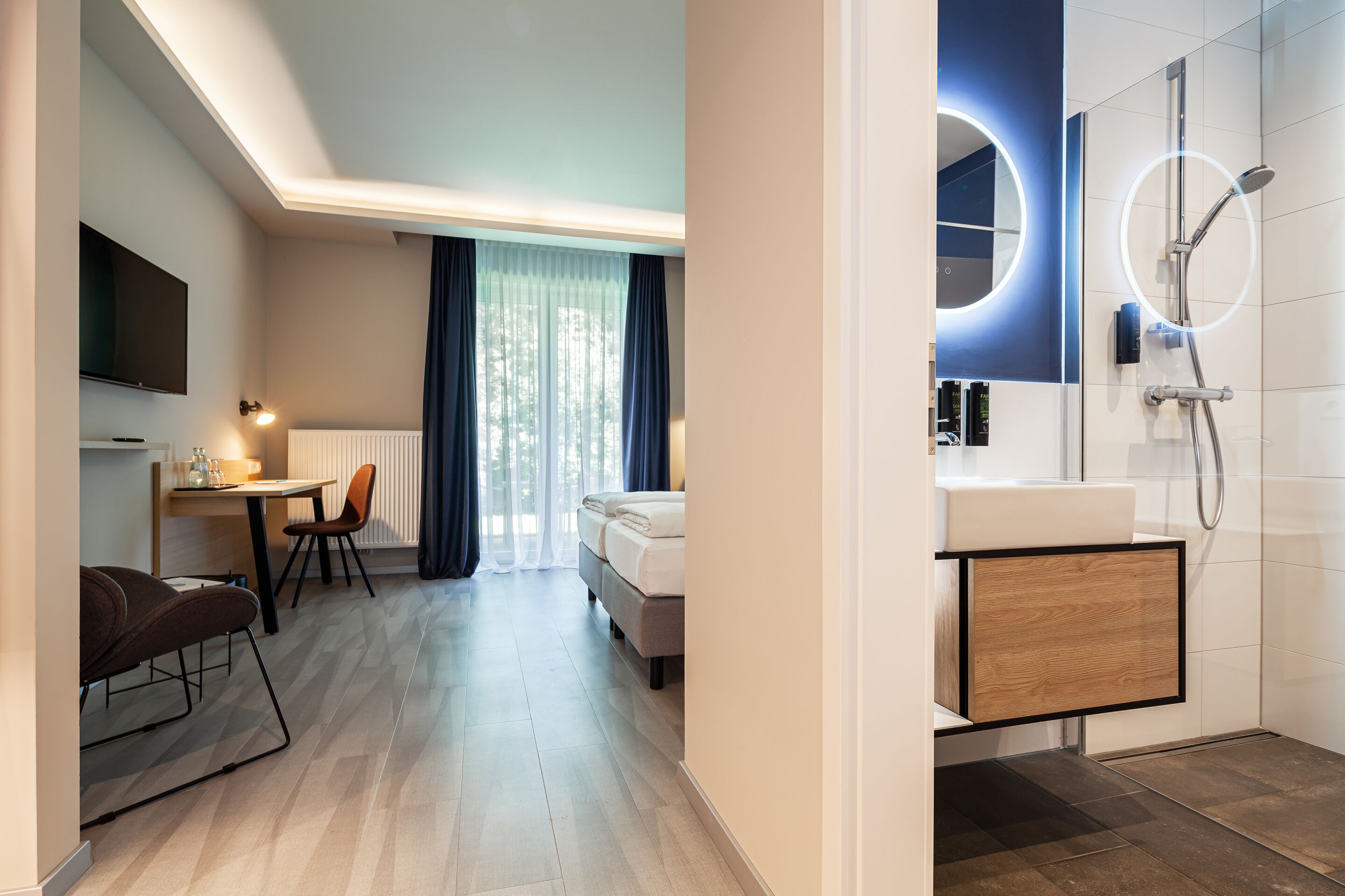 Una habitación de hotel moderna con una cama, un escritorio con silla, televisor y baño con ducha y lavamanos.