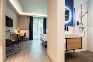 Una habitación de hotel moderna con una cama, un escritorio con silla, televisor y baño con ducha y lavamanos.