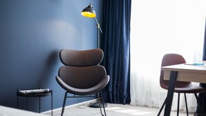 Quarto moderno com parede azul escura, uma mesa redonda, duas cadeiras, um abajur e uma mesinha lateral com uma revista.