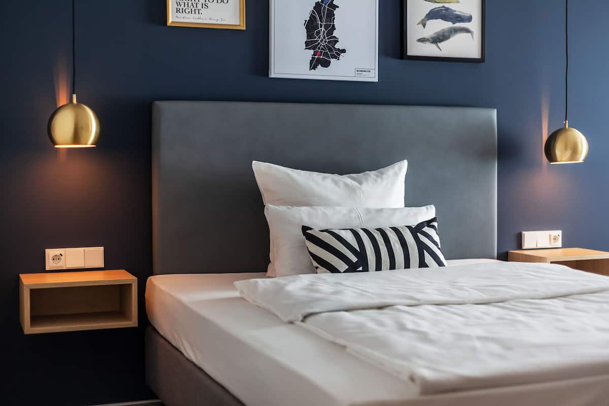 Una cama bien tendida con ropa blanca, un cabecero gris y dos mesitas de noche con lámparas de pared.