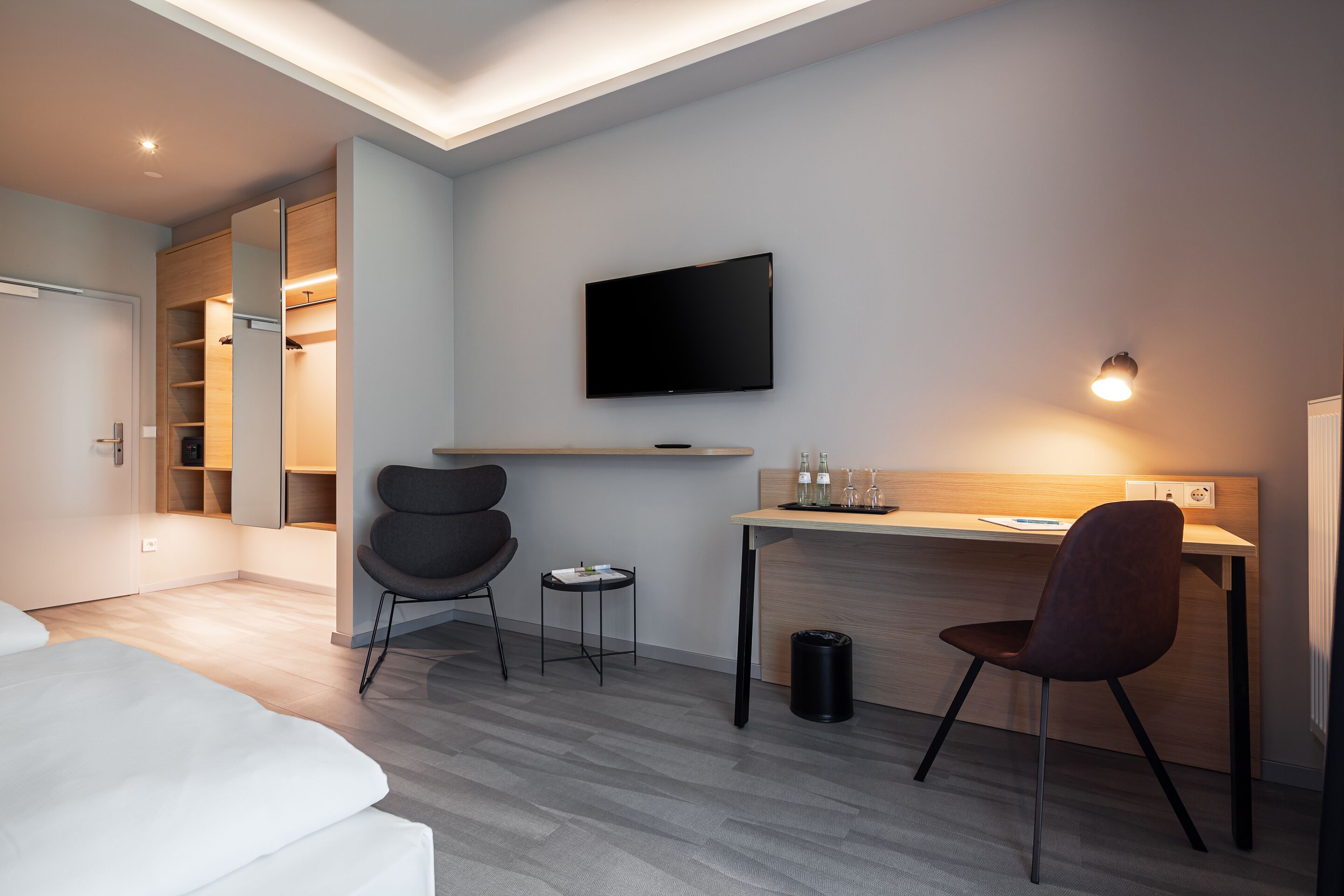 A modern hotel room with a bed, a desk with a chair, a TV, and a wardrobe with a mirror.