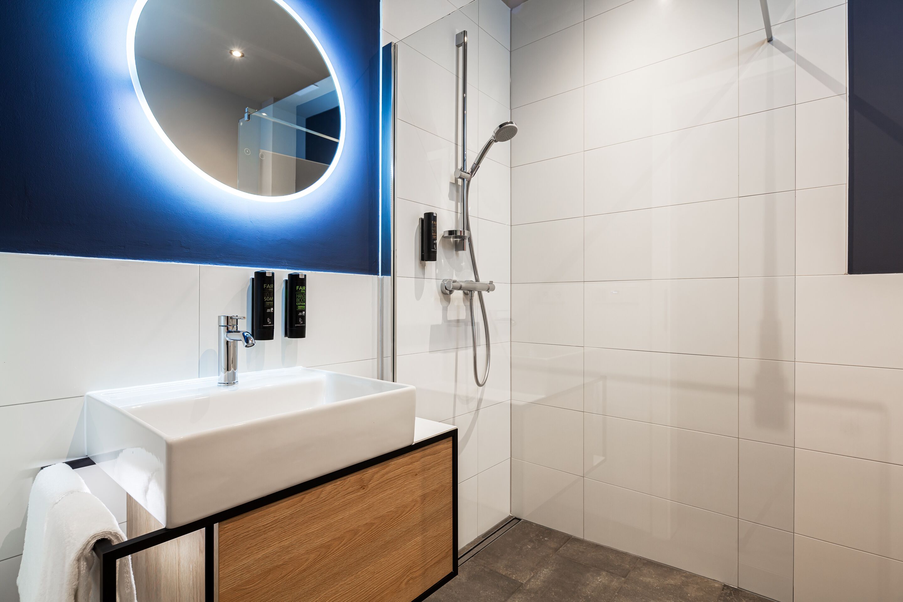 A modern bathroom with a white sink, a wooden cabinet, a mirror with a circular light, and a shower area.