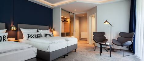 A modern hotel room with two beds, a seating area, and a view of the hallway.