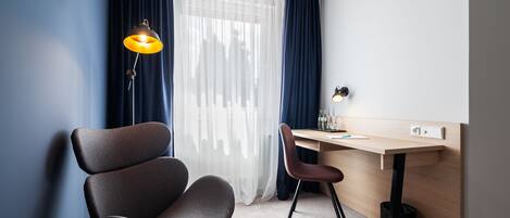 Una habitación de hotel moderna con escritorio, dos sillas, una lámpara y un ventanal con cortinas.
