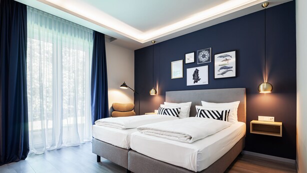 Quarto de hotel moderno com cama de casal, mesas de cabeceira, obra de arte na parede e uma grande janela com cortinas leves.