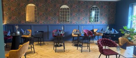 A restaurant with a herringbone wooden floor, round tables with chairs, and a floral wallpaper design.
