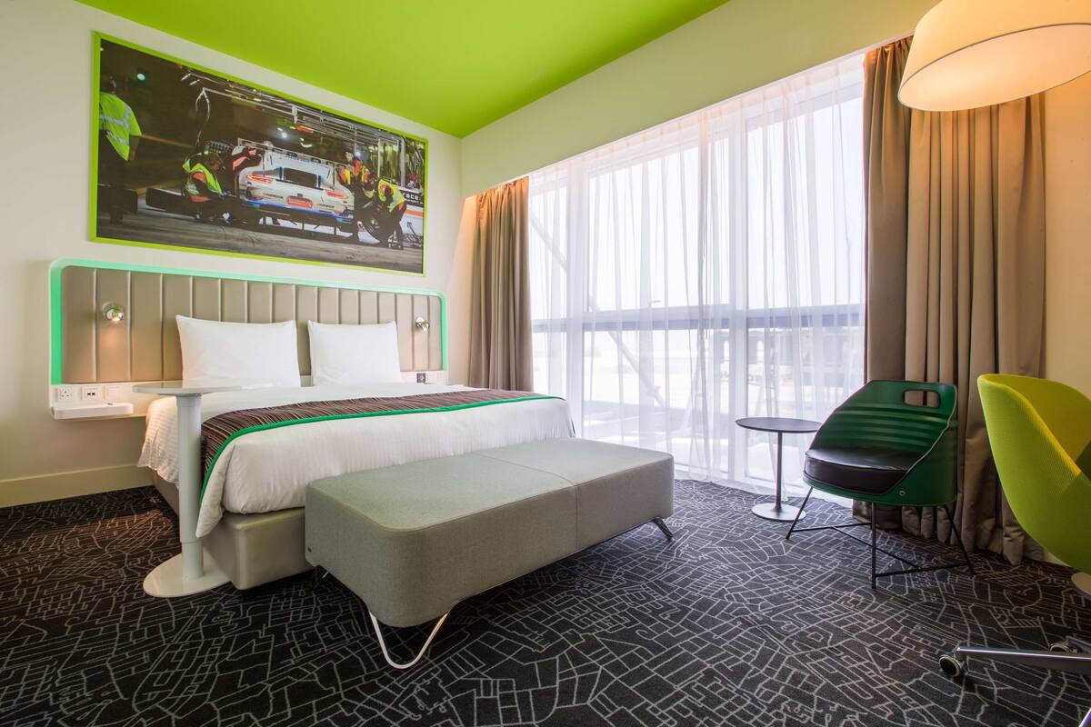 A hotel room with a bed, a green chair, a small table, and a large window with curtains.