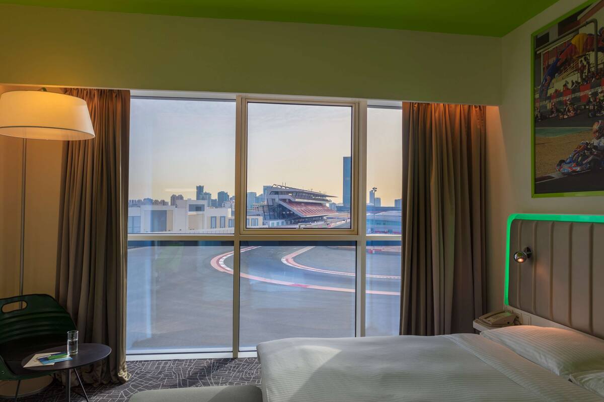 A hotel room with a large window, a bed, a bedside table with a lamp, and a chair.