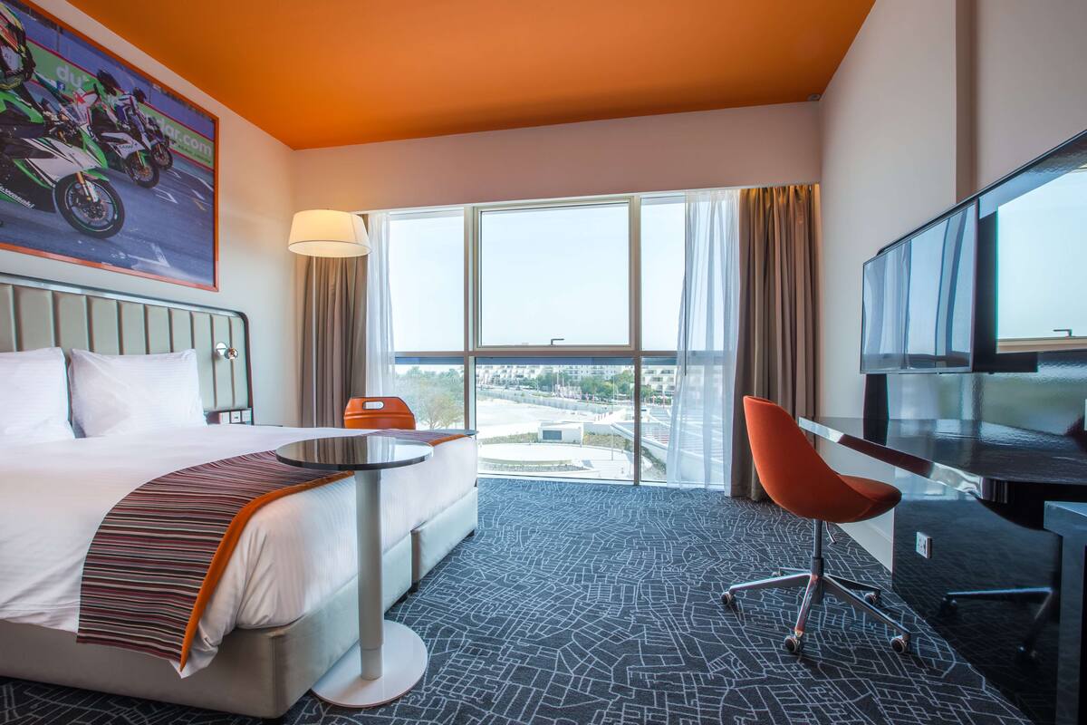 A hotel room with a bed, a desk, a chair, a TV, and large windows offering a view.
