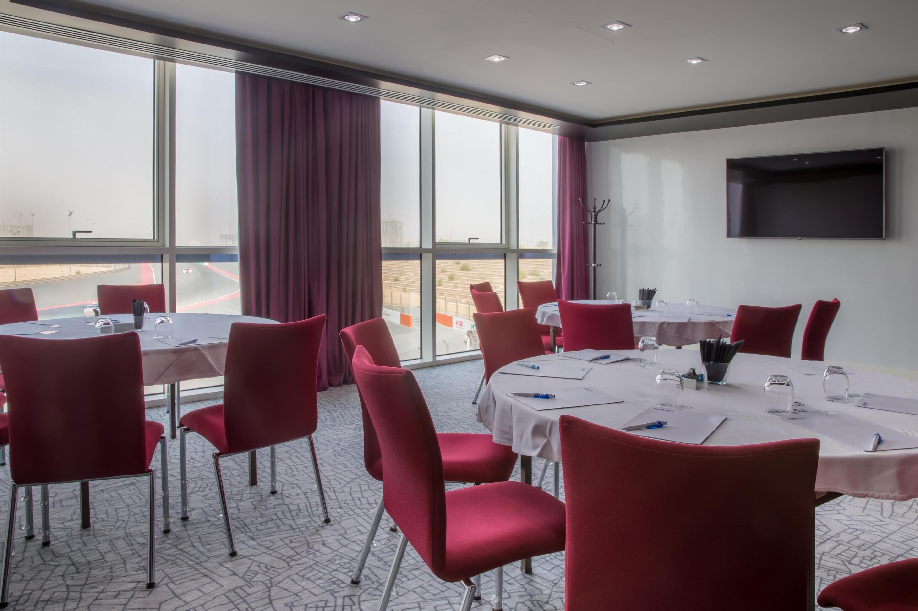 Un salón de conferencias con sillas y mesas rojas, ventanas amplias con cortinas y un televisor de pantalla plana en la pared.