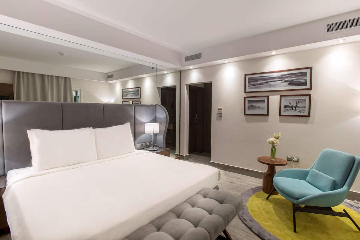 A modern hotel room with a large bed, a grey upholstered headboard, a blue armchair, and a small round table with a vase of flowers.
