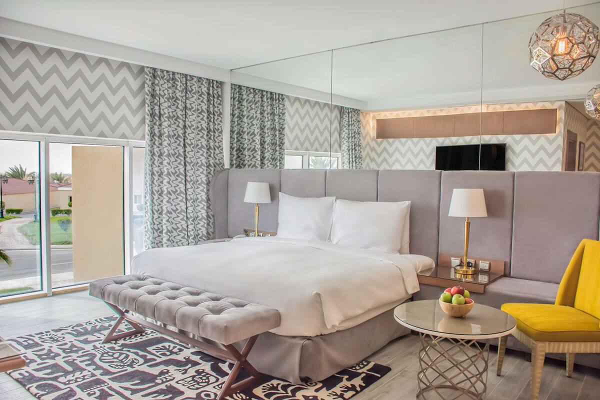 A modern hotel room with a large bed, a sofa, a small table with fruit, and a view of the outdoors.