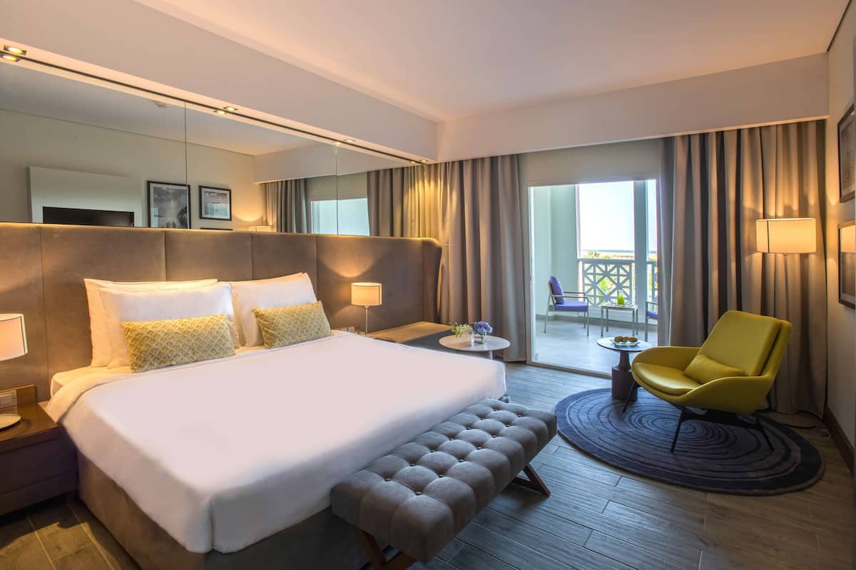 A modern hotel room with a large bed, a seating area with a yellow armchair, and a balcony with a view.