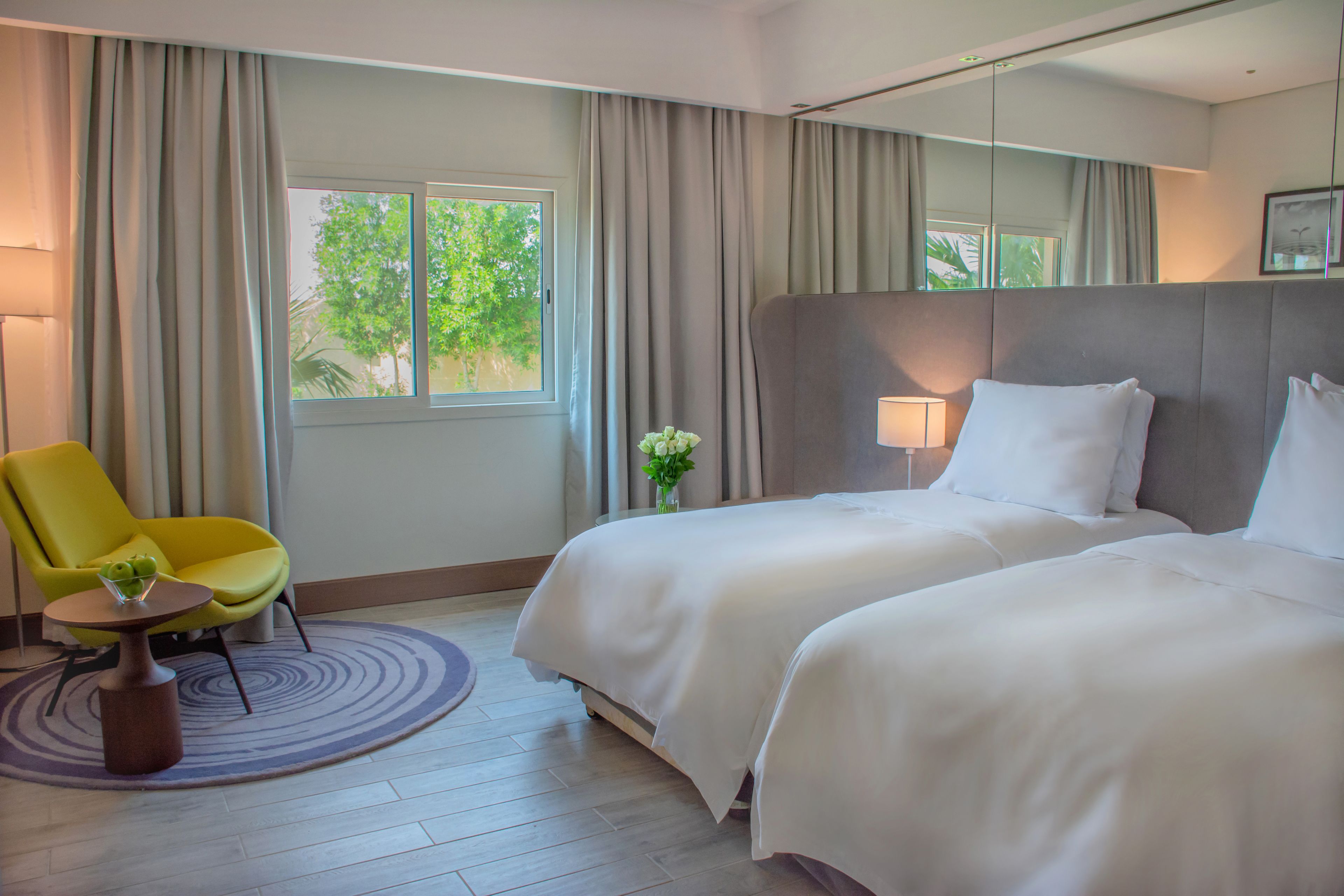 Une chambre d’hôtel avec deux lits, une chaise jaune, une petite table avec un vase et une fenêtre avec des rideaux.