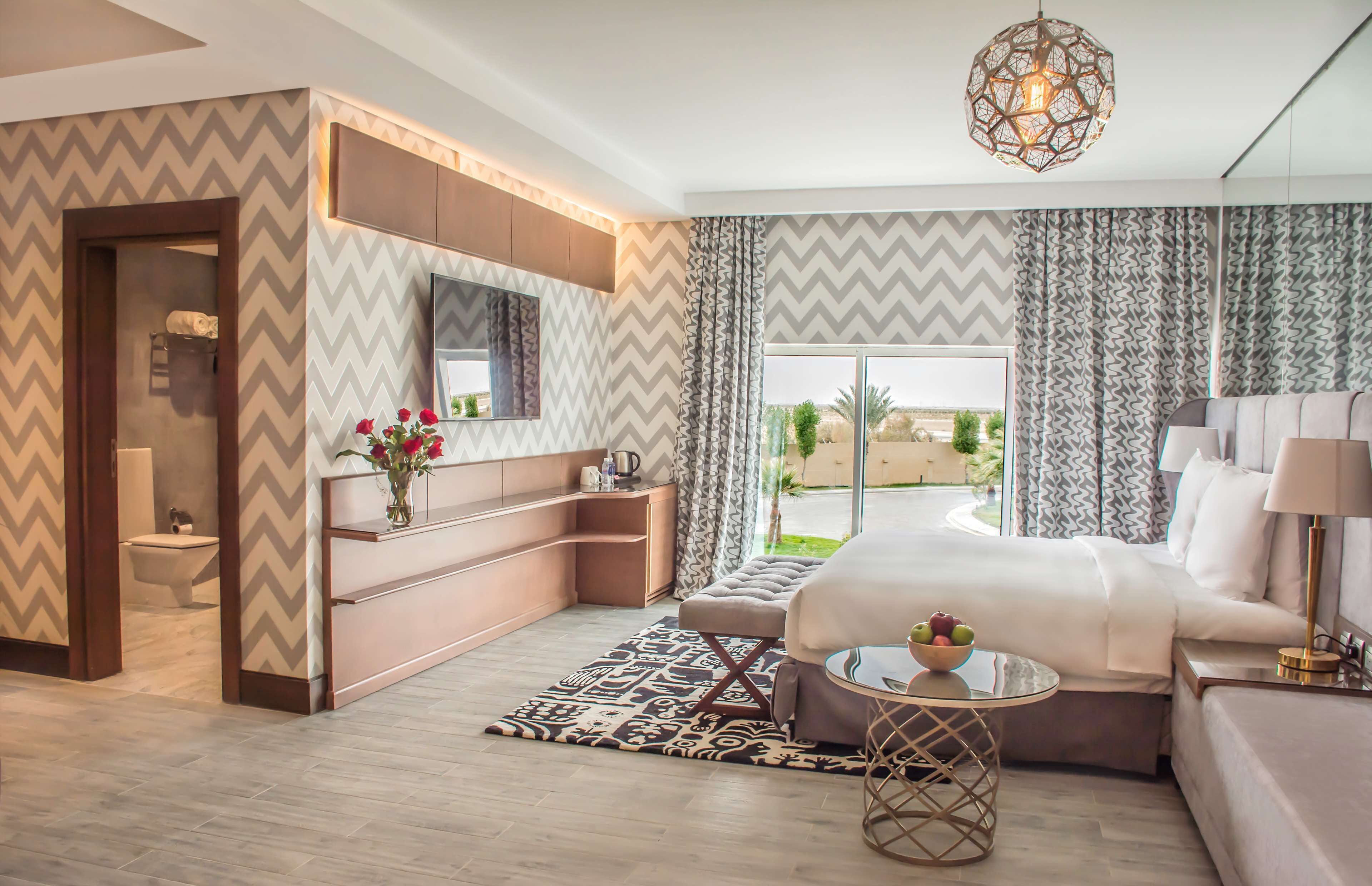 A modern hotel room with a large bed, a sitting area, a vanity, and a view of the outdoors.
