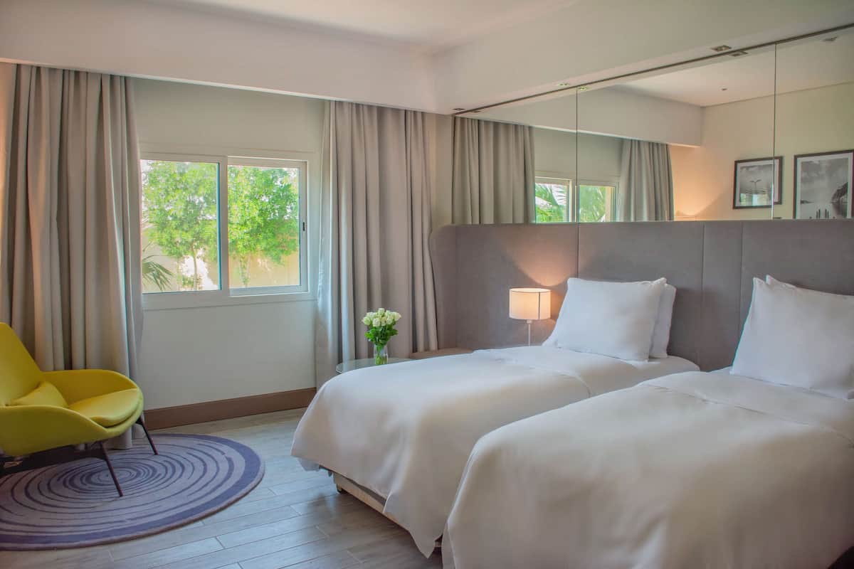 A hotel room with two beds, a yellow chair, a round rug, and a window with curtains.